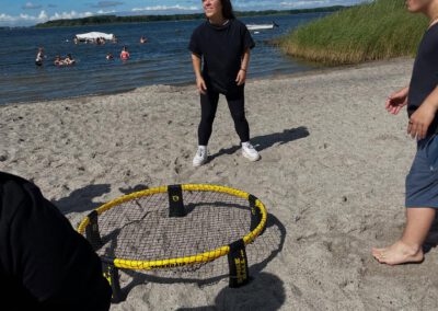 Mehrere Personen spielen am Strand ein Spiel mit einem kleinen runden Netz auf sandigem Boden, im Hintergrund Wasser und Schilf