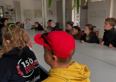Gruppe von Menschen sitzt an einem langen Tisch in einem hellen Raum mit großen Fenstern und Pflanzen im Vordergrund