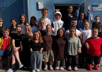 Gruppenfoto von etwa 25 Jugendlichen und jungen Erwachsenen vor einem blau-weißen Gebäude. Die Gruppe lächelt und posiert in die Kamera. Sonniges Wetter. Viele lachen und schauen in die Kamera; einzelne winken oder strecken die Hände hoch.