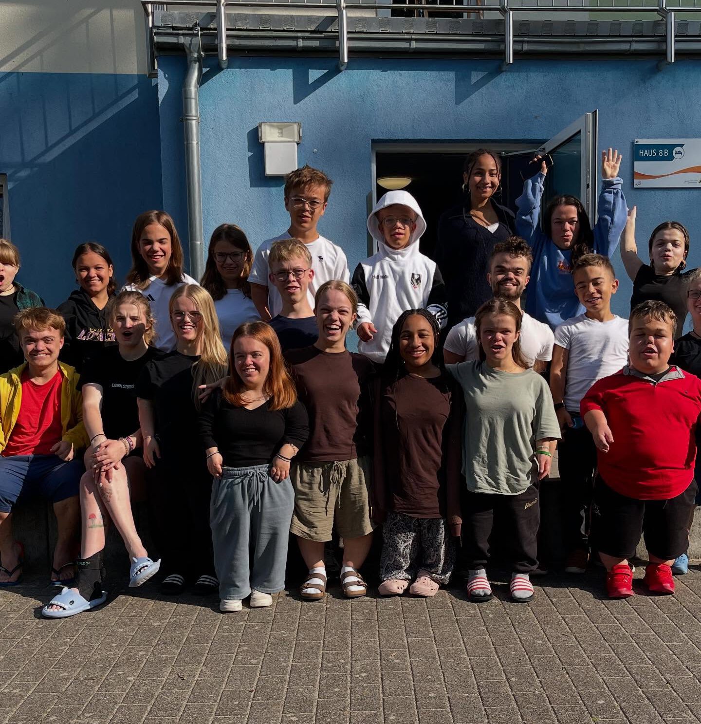 Gruppenfoto von etwa 25 Jugendlichen und jungen Erwachsenen vor einem blau-weißen Gebäude. Die Gruppe lächelt und posiert in die Kamera. Sonniges Wetter. Viele lachen und schauen in die Kamera; einzelne winken oder strecken die Hände hoch.