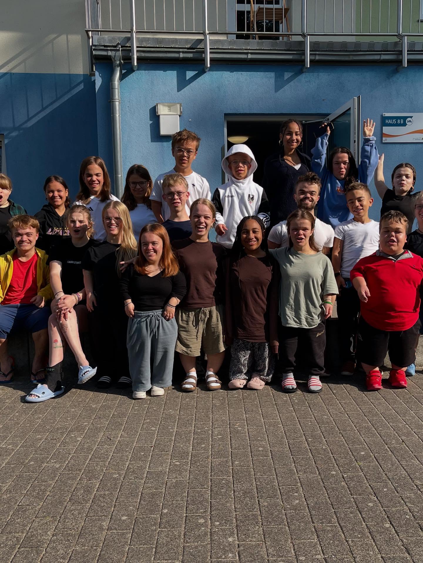 Gruppenfoto von etwa 25 Jugendlichen und jungen Erwachsenen vor einem blau-weißen Gebäude. Die Gruppe lächelt und posiert in die Kamera. Sonniges Wetter. Viele lachen und schauen in die Kamera; einzelne winken oder strecken die Hände hoch.
