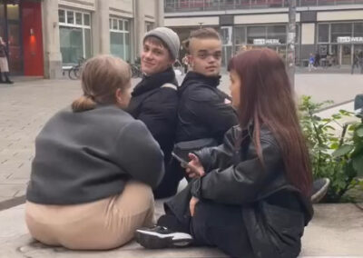 vier Jugendliche Sitzen auf einer Bank und unterhalten sich, dahinter ein Bahnhof