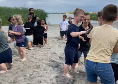 Eine Gruppe von Jugendlichen und Betreuern spielen gemeinsam ein Laufspiel am Sandstrand eines Sees unter bewölktem Himmel