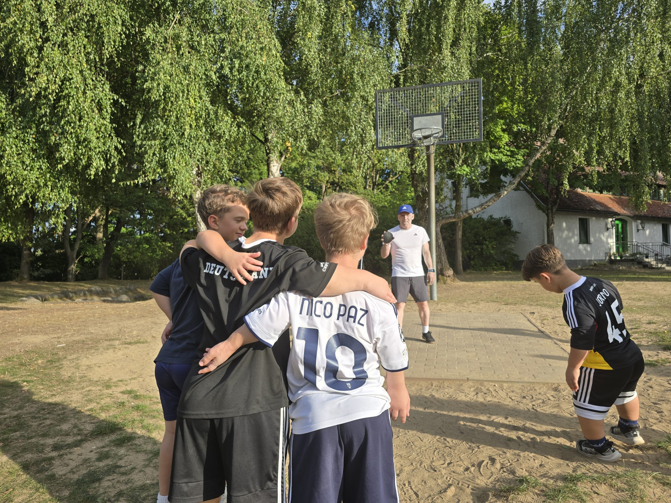 20260912_172722 Drei Kinder in Fußballtrikots stehen Arm in Arm mit dem Rücken zur Kamera auf einem Sandplatz, dahinter hält ein Erwachsener einen Diskus vor einem Basketballkorb.