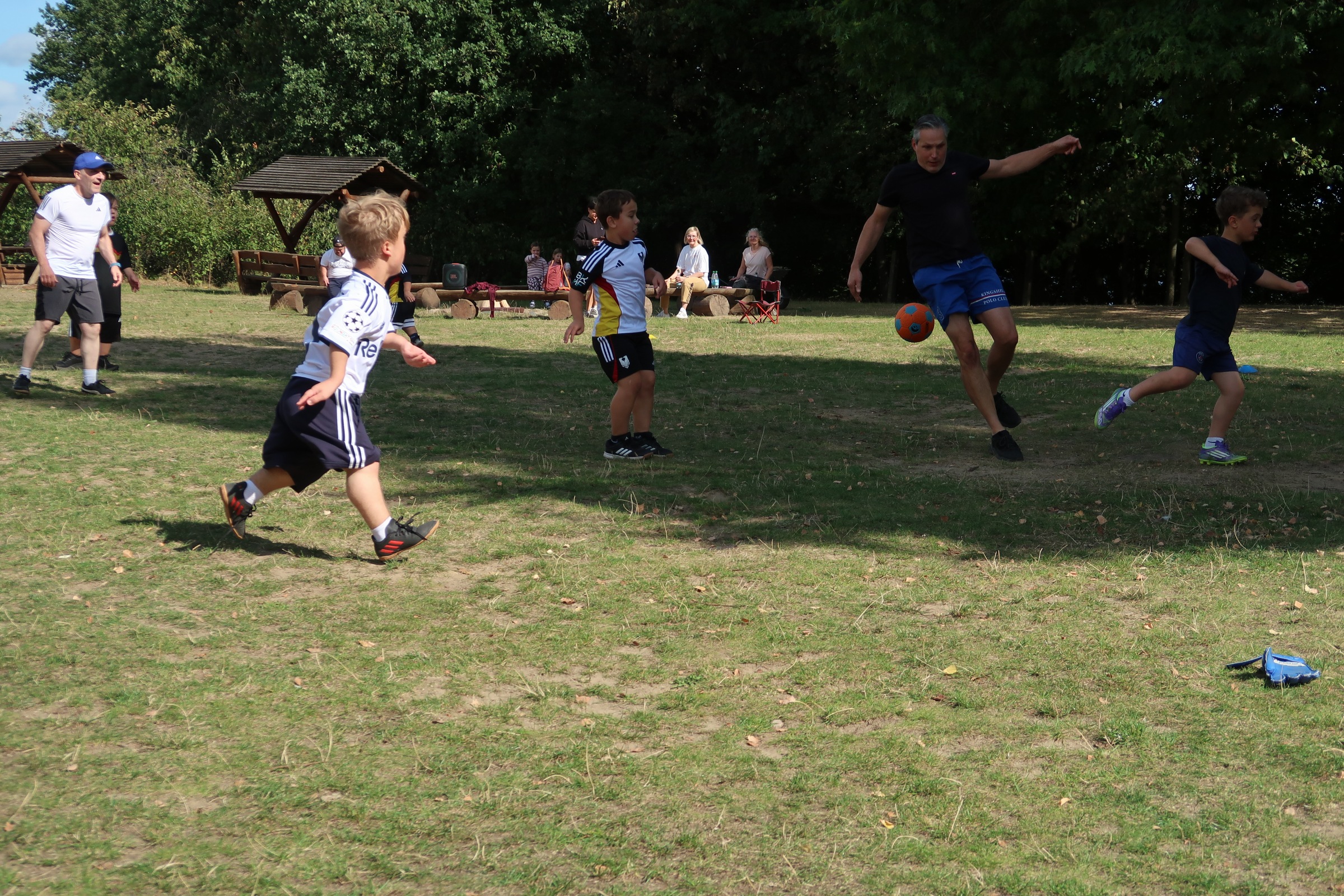 IMG_2355 Kinder und ein Erwachsener kämpfen auf einer Wiese um einen orangefarbenen Fußball, im Hintergrund schauen Zuschauende von Baumstammbänken zu.
