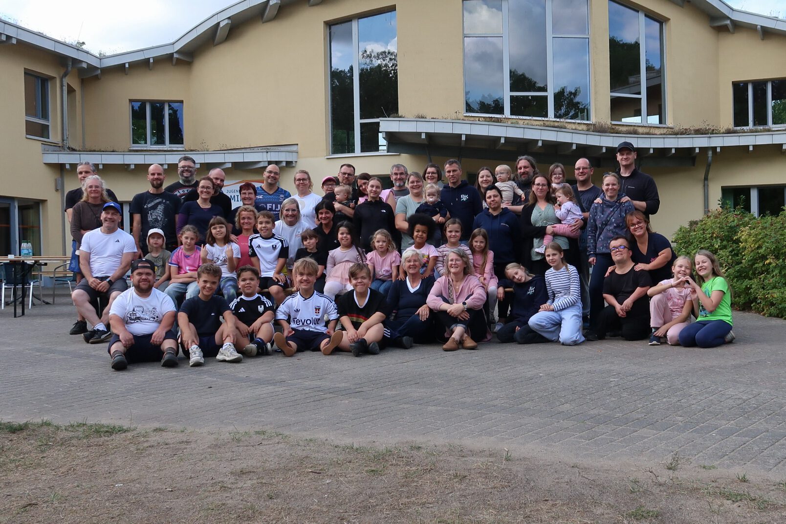 Gruppenfoto der Teilnehmenden vor einem gelben Gebäude mit geschwungenem Dach: Erwachsene stehen hinten, Kinder sitzen vorn auf dem Pflaster.