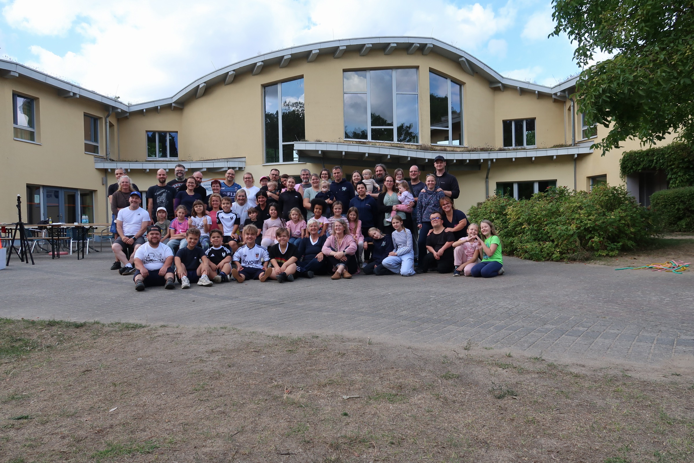 Gruppenfoto der Teilnehmenden vor einem gelben Gebäude mit geschwungenem Dach: Erwachsene stehen hinten, Kinder sitzen vorn auf dem Pflaster.