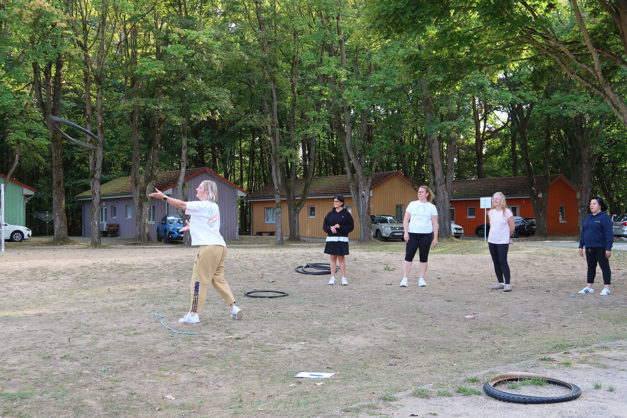 IMG_2435 Fünf Frauen stehen auf einer Wiese zwischen Bäumen, eine wirft einen Reifen, die anderen schauen zu; dahinter bunte Holzhütten.