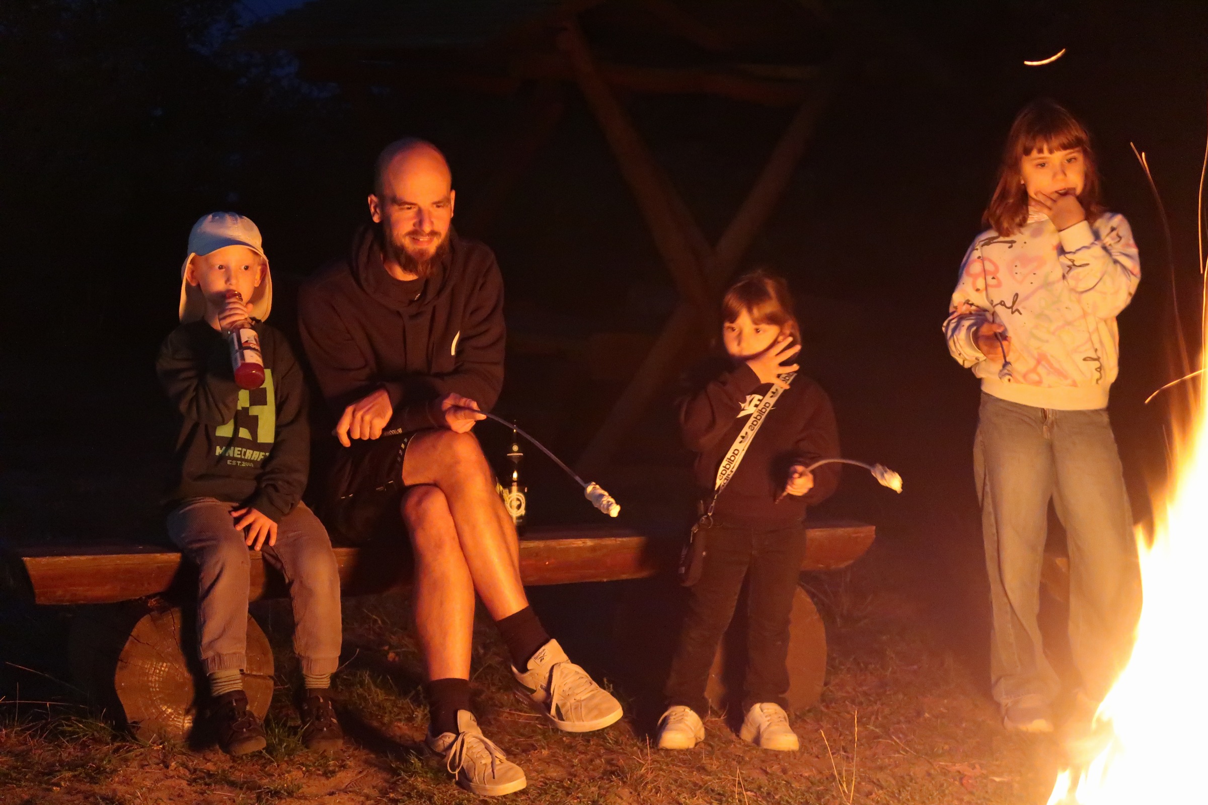 IMG_2459 Abends am Lagerfeuer: Ein Mann mit Bart sitzt auf einer Holzbank, drei Kinder daneben halten Stöcke mit Stockbrot ins Feuer.