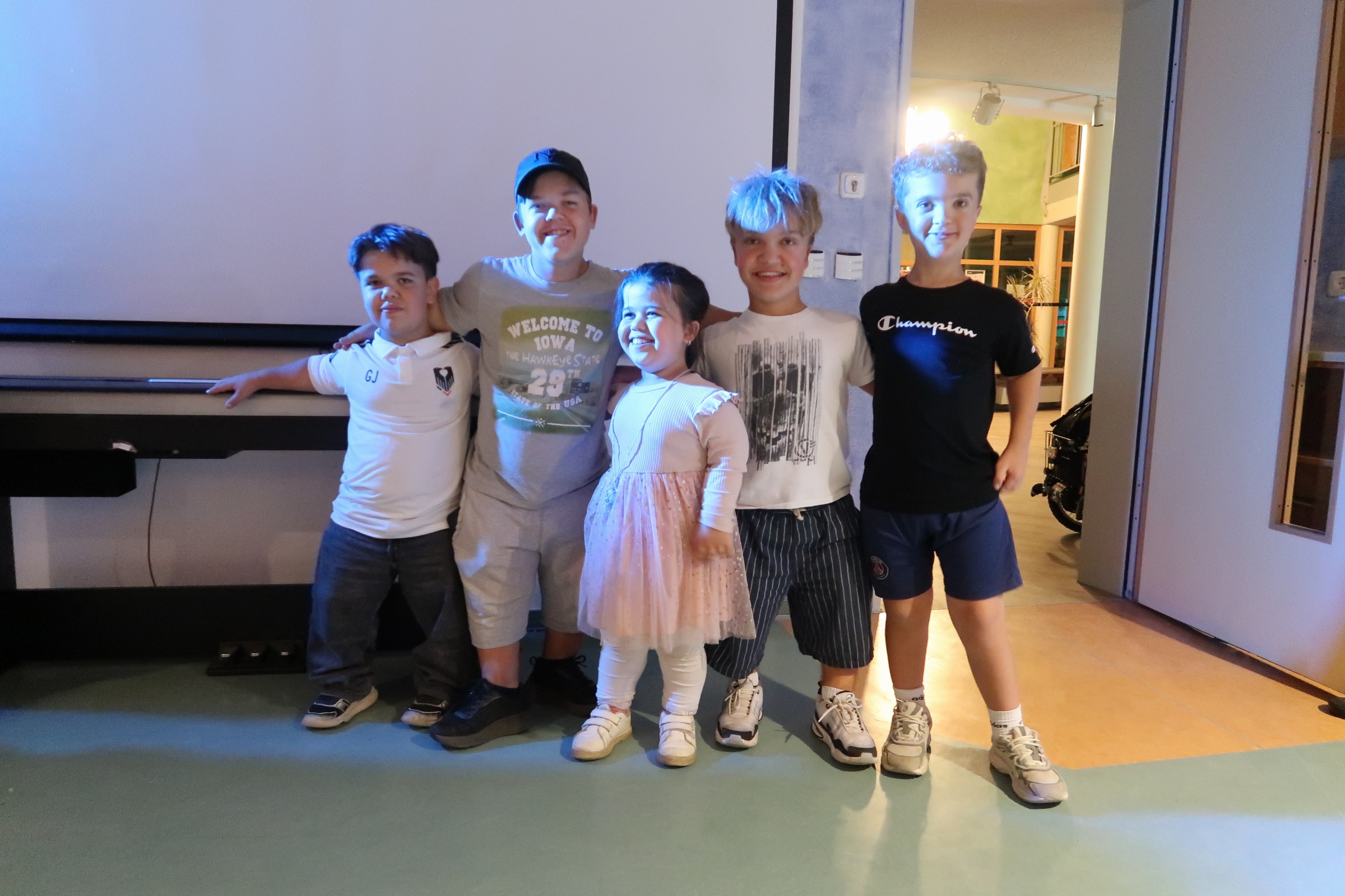 IMG_2478 Fünf lächelnde Kinder und Jugendliche stehen Arm in Arm vor einer Leinwand.