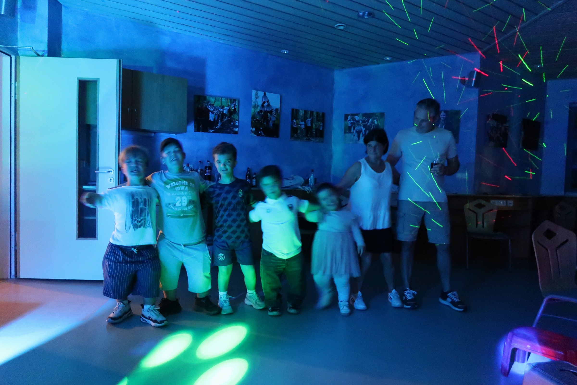 IMG_2491 Fünf Kinder und zwei Erwachsene stehen Arm in Arm in einem blau beleuchteten Raum, grüne und rote Laserstrahlen leuchten an Decke und Wand.